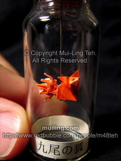 A cute orange origami kyuubi no kitsune (nine-tailed fox), hanging off a needline in a bottle, by Mui-Ling Teh. The yellow label at the bottom says "nine-tailed fox" in Japanese.