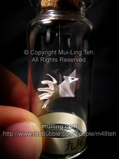 A cute white origami kyuubi no kitsune (nine-tailed fox), hanging off a needline in a bottle, by Mui-Ling Teh. The yellow label at the bottom says "nine-tails" in Japanese.